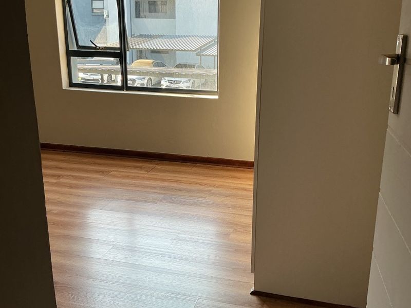 A bright, empty bedroom featuring modern laminate flooring and a large aluminium-framed window with views of the complex parking area.