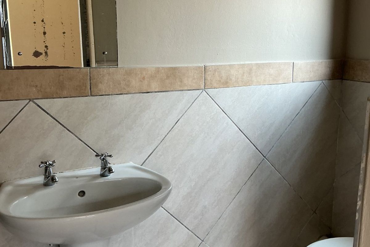 A compact bathroom featuring a pedestal basin, toilet, and neutral ceramic tiling. The space requires minor cosmetic updates to the mirror and finishes.