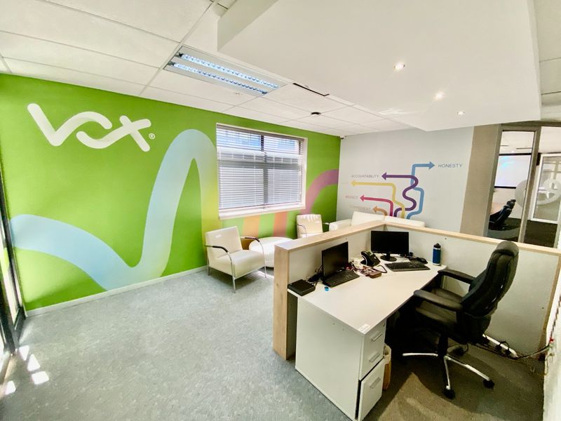 A bright, open-plan office space featuring carpeted flooring, suspended ceilings with recessed lighting, and large windows with venetian blinds.