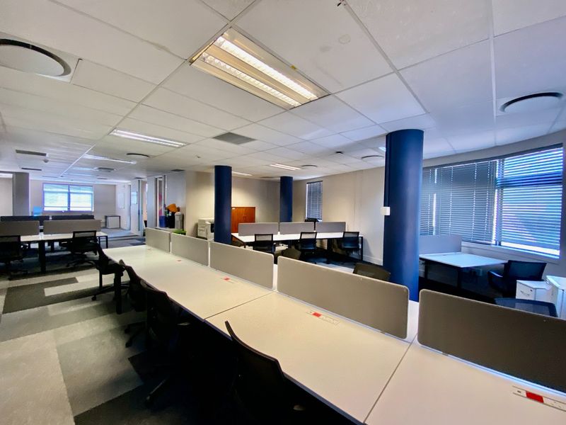 Open-plan commercial office space featuring suspended ceilings, structural columns, carpet tiles, and large windows with venetian blinds.