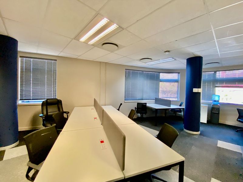 Open-plan commercial office space featuring suspended ceilings, recessed lighting, large windows with venetian blinds, and durable carpet flooring.