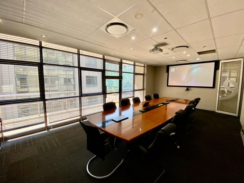 A professional boardroom featuring a large conference table, integrated projection system, suspended ceiling, and expansive windows with venetian blinds.