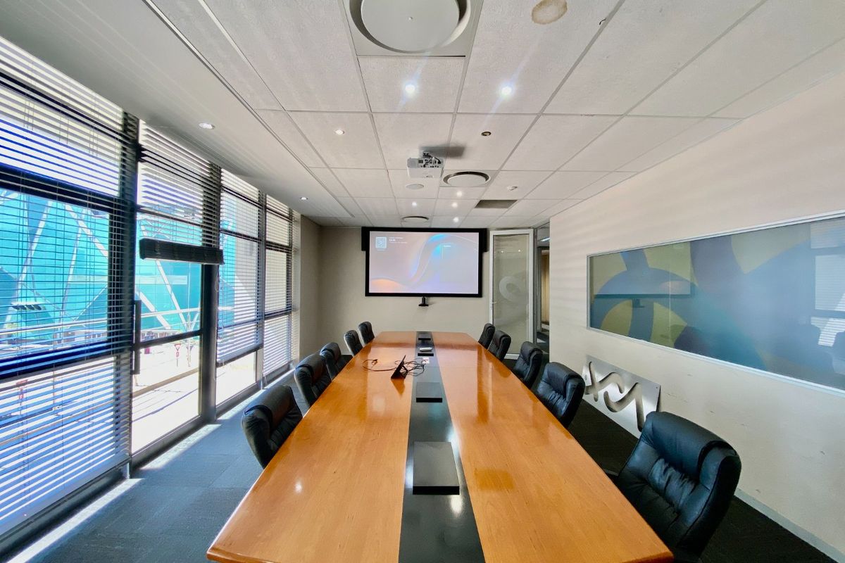 A professional boardroom featuring a large conference table, integrated projector screen, suspended ceiling with recessed lighting, and floor-to-ceiling windows with venetian blinds.