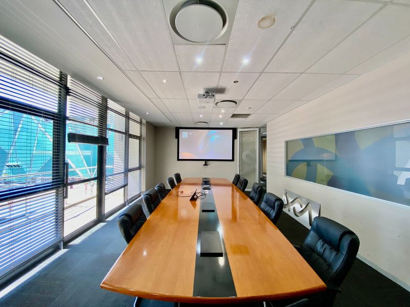 A professional boardroom featuring a large conference table, integrated projector screen, suspended ceiling with recessed lighting, and floor-to-ceiling windows with venetian blinds.