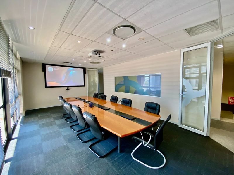 A professional boardroom featuring a large conference table, integrated projector system, suspended ceiling, and glass partition walls for a modern commercial workspace.