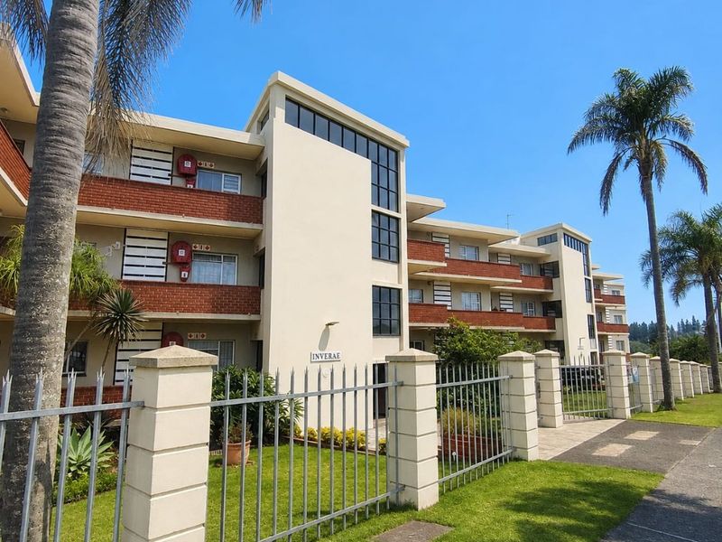 Front exterior view of the Inverae apartment complex featuring brick and concrete facade, metal perimeter fencing, and well-maintained lawn.