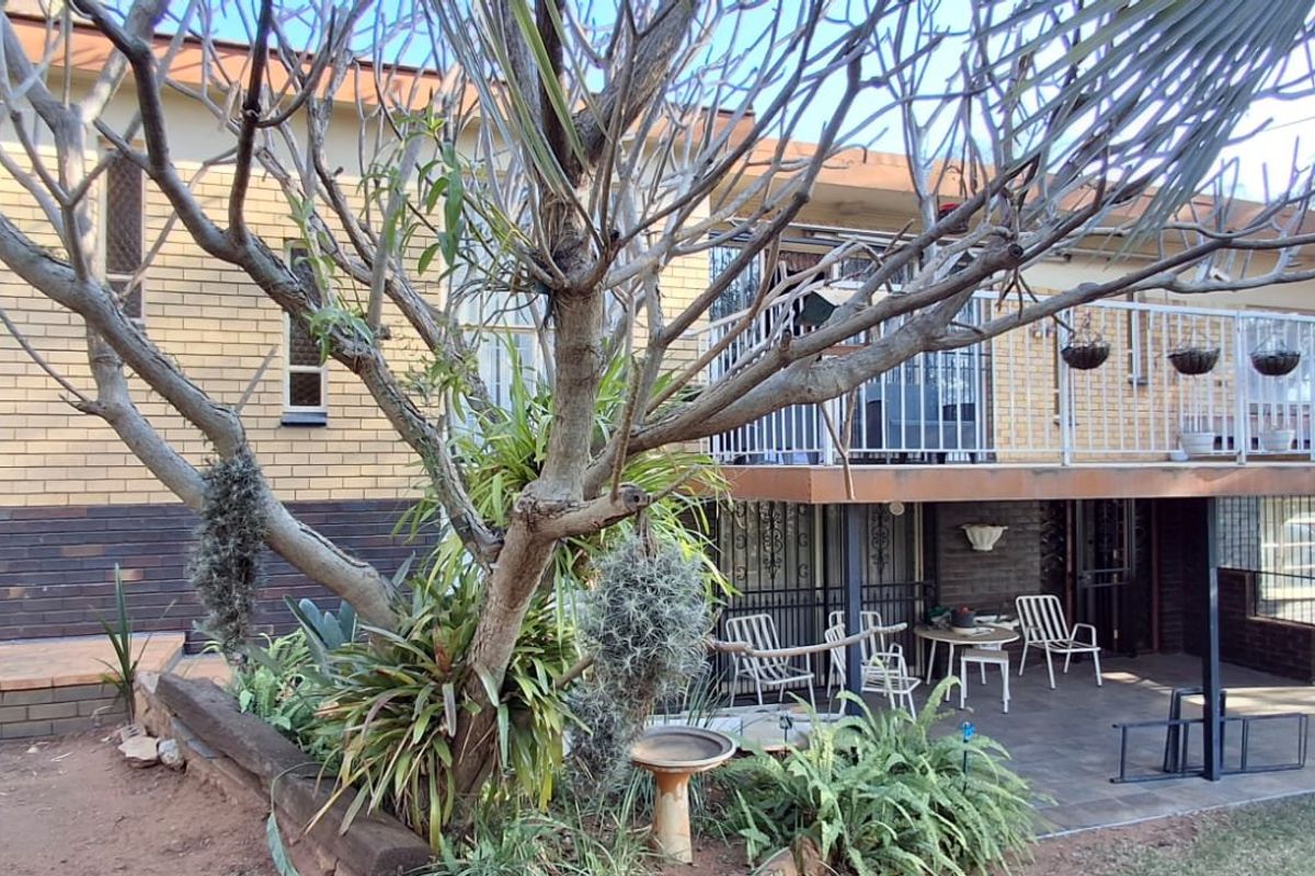 Rear view of a multi-level brick house featuring a covered patio area, an upper-level balcony with metal railings, and established garden landscaping.