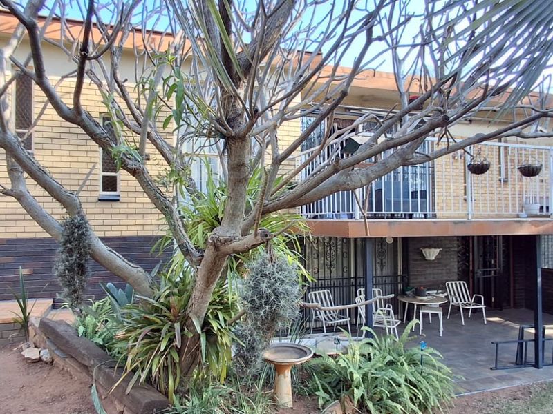 Rear view of a multi-level brick house featuring a covered patio area, an upper-level balcony with metal railings, and established garden landscaping.