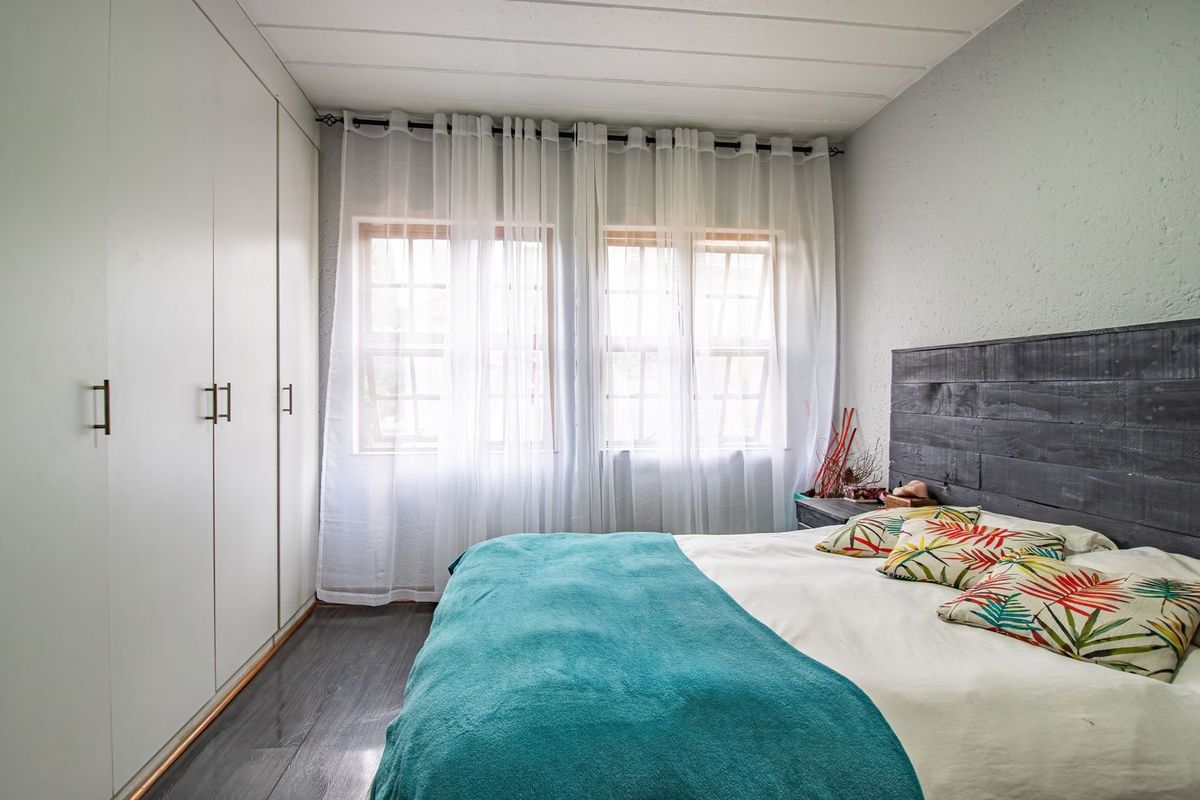 A well-lit bedroom featuring extensive built-in cupboards, dark laminate flooring, and large timber-framed windows with sheer curtains.