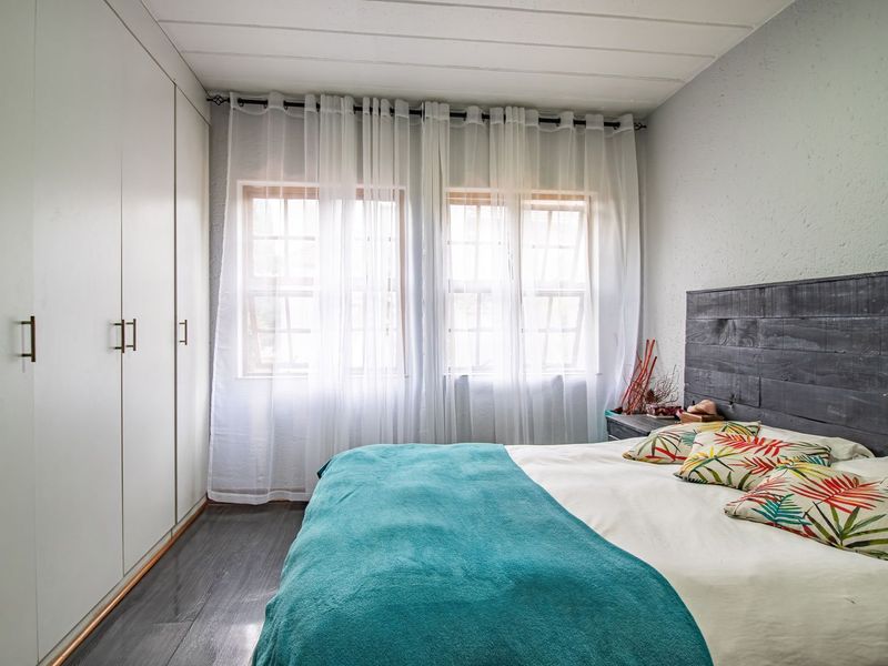 A well-lit bedroom featuring extensive built-in cupboards, dark laminate flooring, and large timber-framed windows with sheer curtains.