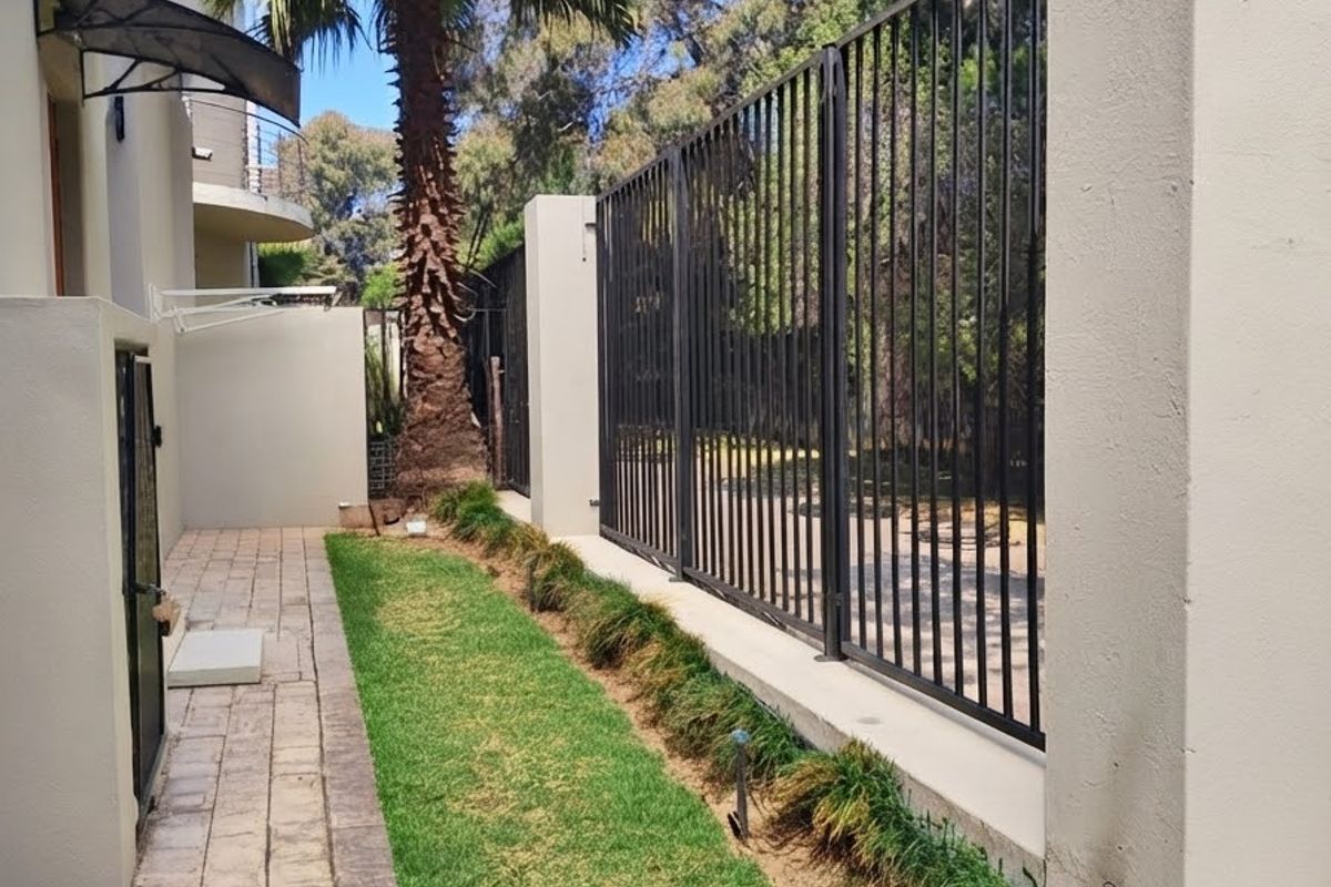 A side garden area featuring a paved walkway, manicured lawn, mature palm tree, and secure metal perimeter fencing.