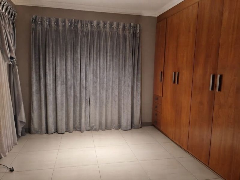 A spacious bedroom featuring neutral tiled flooring, extensive built-in wooden cabinetry, and floor-to-ceiling curtains.