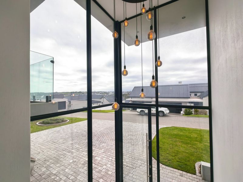 Modern entrance featuring floor-to-ceiling steel-framed windows, a glass door, and a striking pendant light cluster overlooking the paved driveway and neighbourhood.