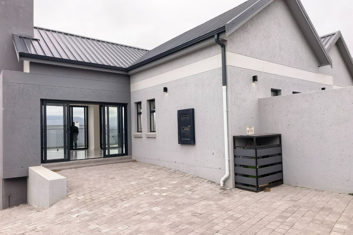 A modern rear exterior view featuring a paved courtyard, sliding glass doors, and a well-maintained grey rendered facade with a metal roof.