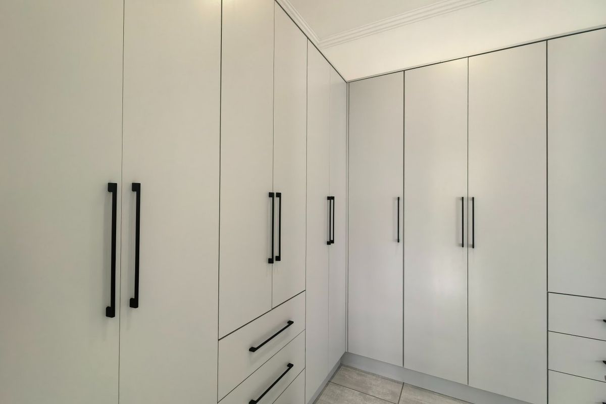 A clean, modern bedroom featuring extensive floor-to-ceiling built-in cupboards with matte black hardware and neutral tiled flooring.