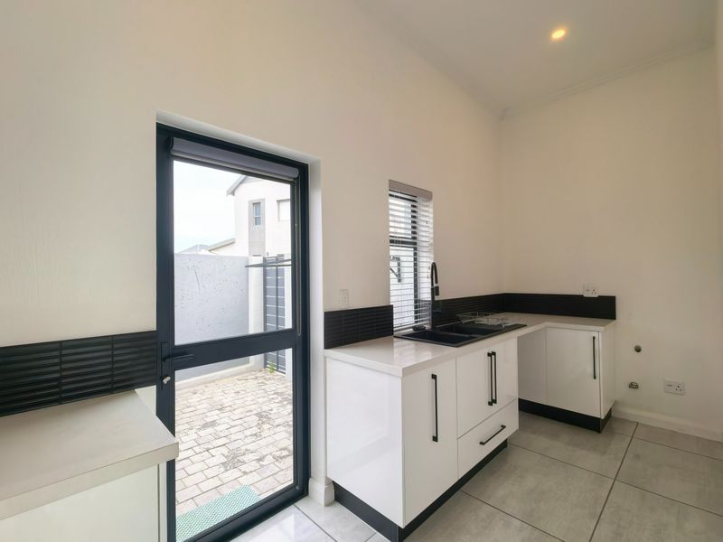 A modern, clean scullery featuring white cabinetry, black tiled splashbacks, stone countertops, and direct access to an outdoor paved area.