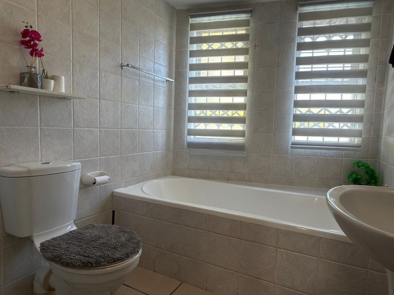 A functional bathroom featuring a built-in bathtub, pedestal sink, and neutral ceramic tiling throughout, complemented by dual windows with fitted blinds.