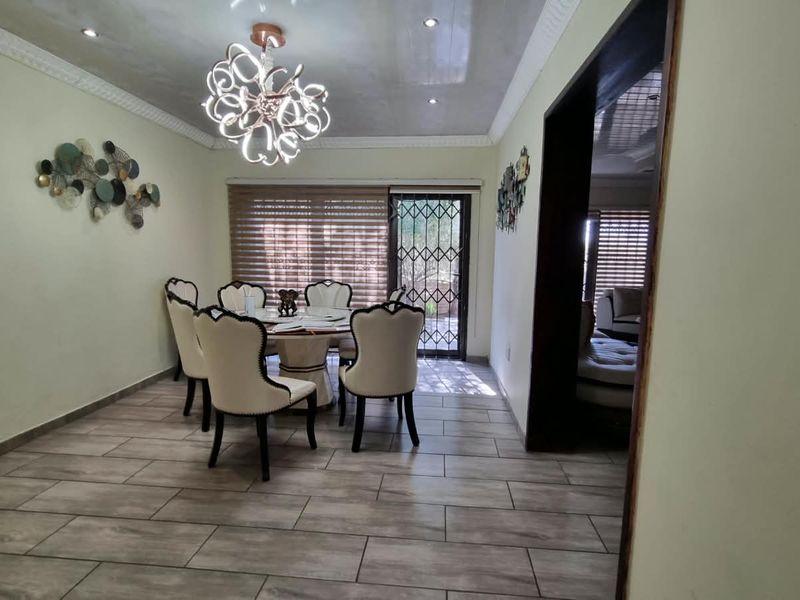 A well-lit dining room featuring modern tiled flooring, decorative cornicing, and a stylish chandelier, with direct access to an outdoor area via a security gate.
