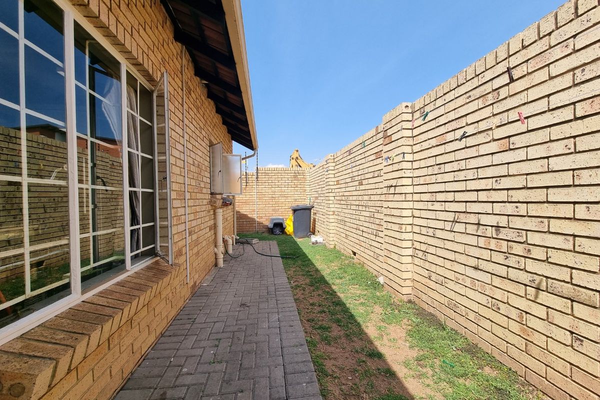 A side exterior view of the townhouse featuring a paved walkway, face brick facade, and a high boundary wall.