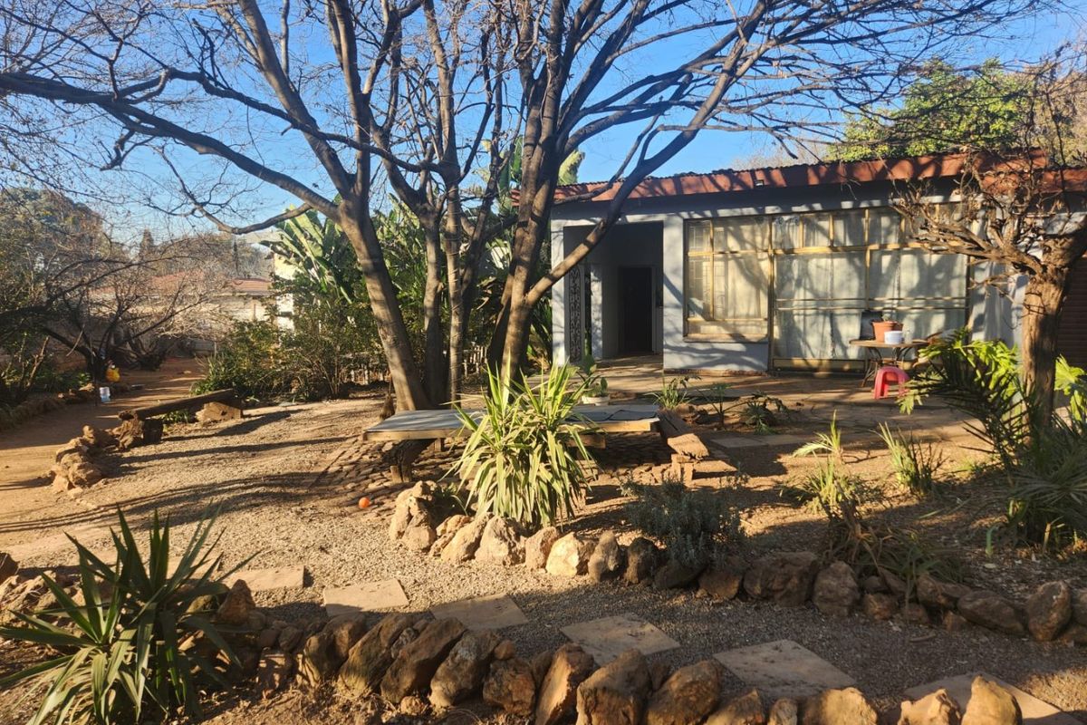 A rear garden view featuring mature trees, stone landscaping, and a covered patio area attached to the main house structure.