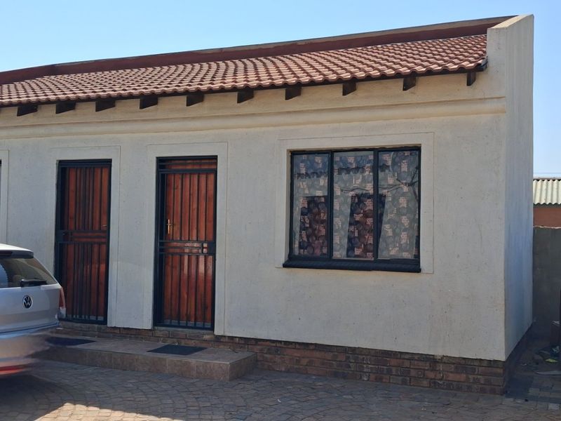 Front elevation of a single-story residential house featuring a terracotta tiled roof, security gates, and a paved driveway area.