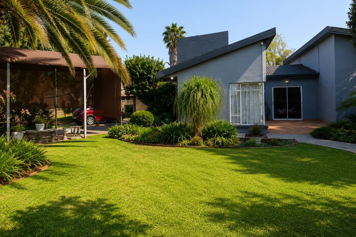 A well-maintained rear garden featuring a lush lawn, landscaped garden beds, a paved walkway, and a covered carport area adjacent to the home.