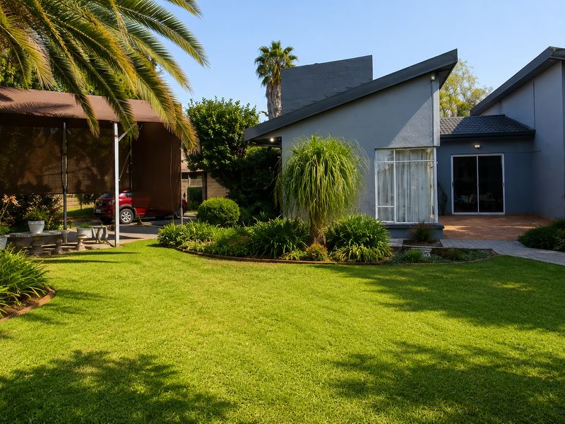 A well-maintained rear garden featuring a lush lawn, landscaped garden beds, a paved walkway, and a covered carport area adjacent to the home.
