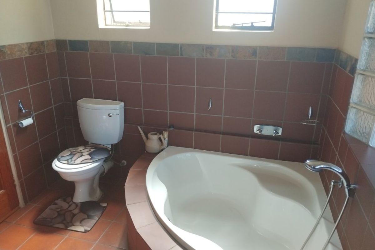 A functional bathroom featuring a corner bathtub, toilet, and tiled finishes with natural light from louver and glass block windows.