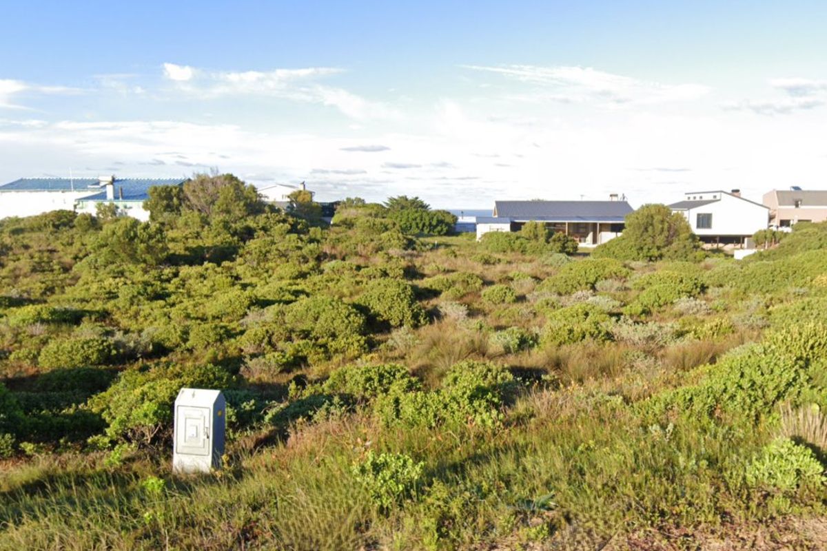 A vacant residential plot covered in coastal vegetation with a visible electrical utility box, situated in a developed neighborhood.