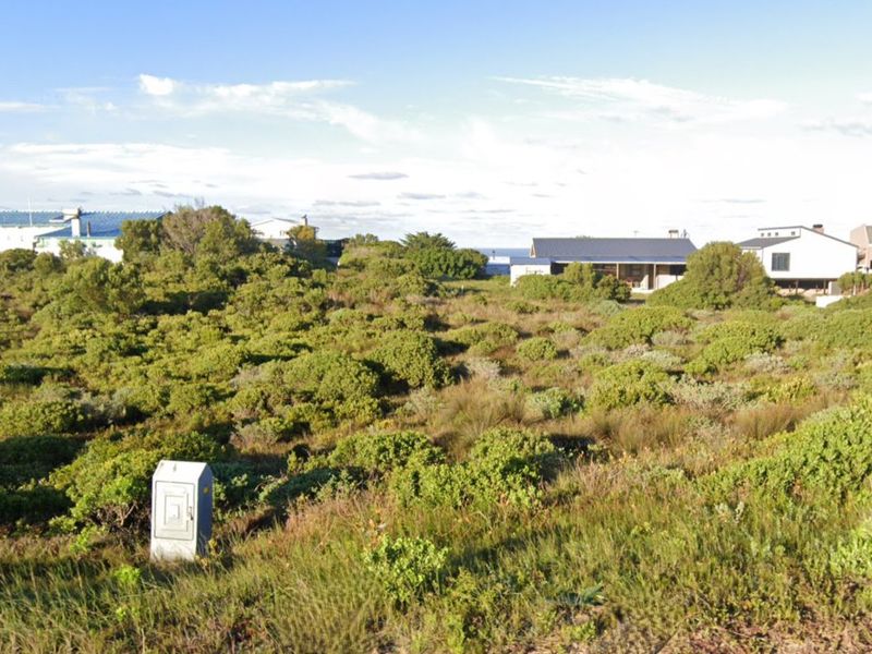 A vacant residential plot covered in coastal vegetation with a visible electrical utility box, situated in a developed neighborhood.