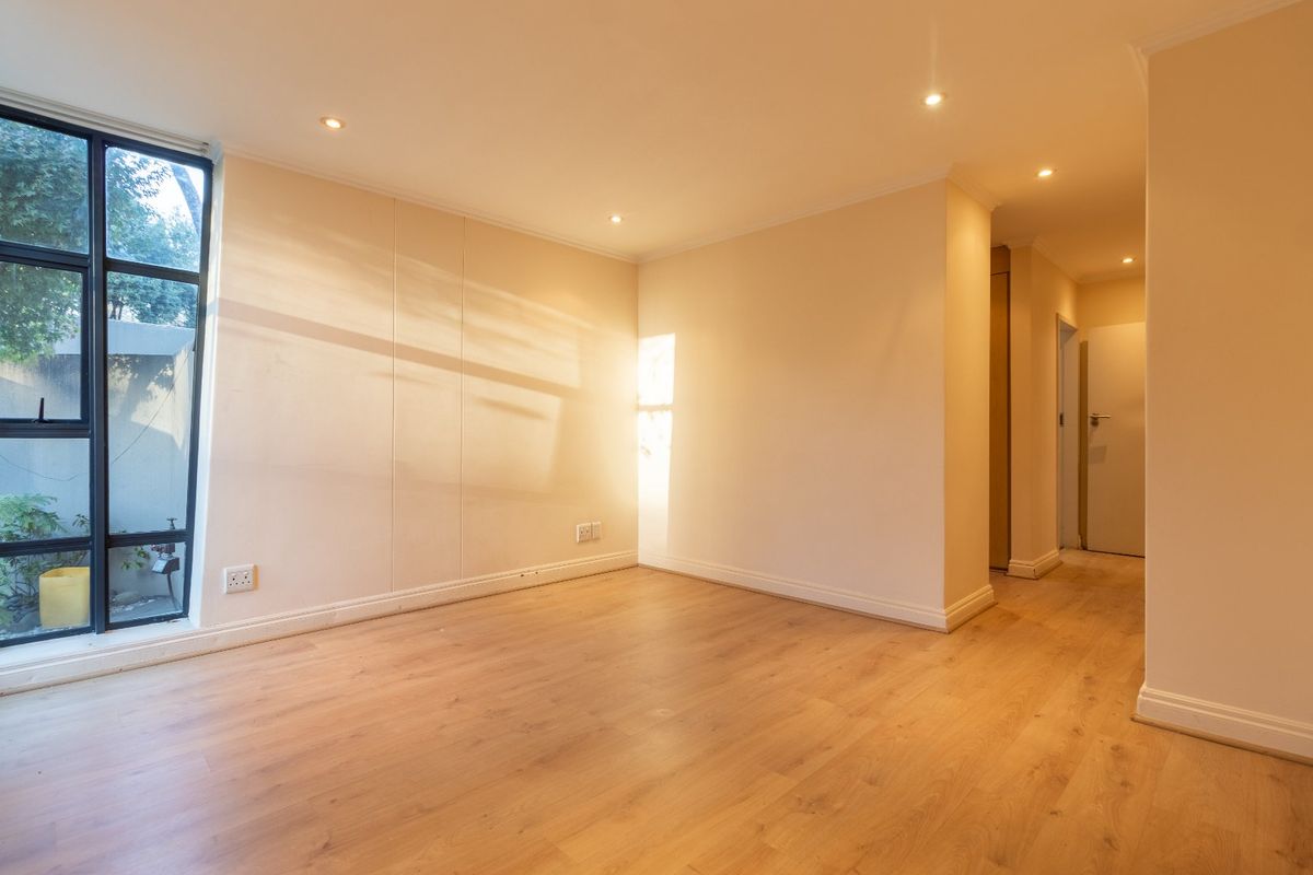 A bright, empty living area featuring modern laminate flooring, recessed downlighting, and a large window with views of the exterior.