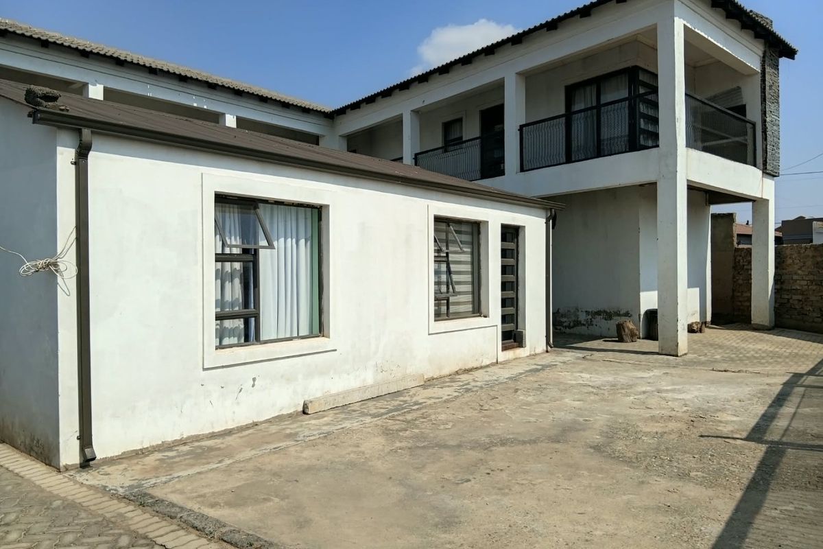 Rear exterior view of a two-story house featuring a paved courtyard, upper-level balcony with metal railing, and white rendered walls.