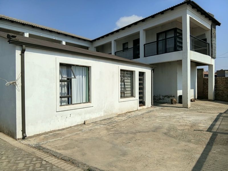 Rear exterior view of a two-story house featuring a paved courtyard, upper-level balcony with metal railing, and white rendered walls.