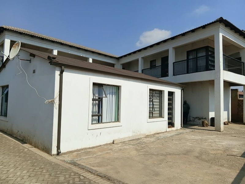 Rear view of a two-story residential building featuring a paved courtyard, balcony with metal railings, and white exterior walls.