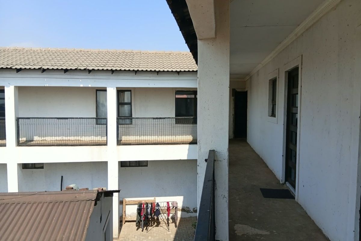 An exterior walkway balcony featuring metal railings and concrete flooring, showing signs of wear and requiring maintenance to the exterior wall finishes.