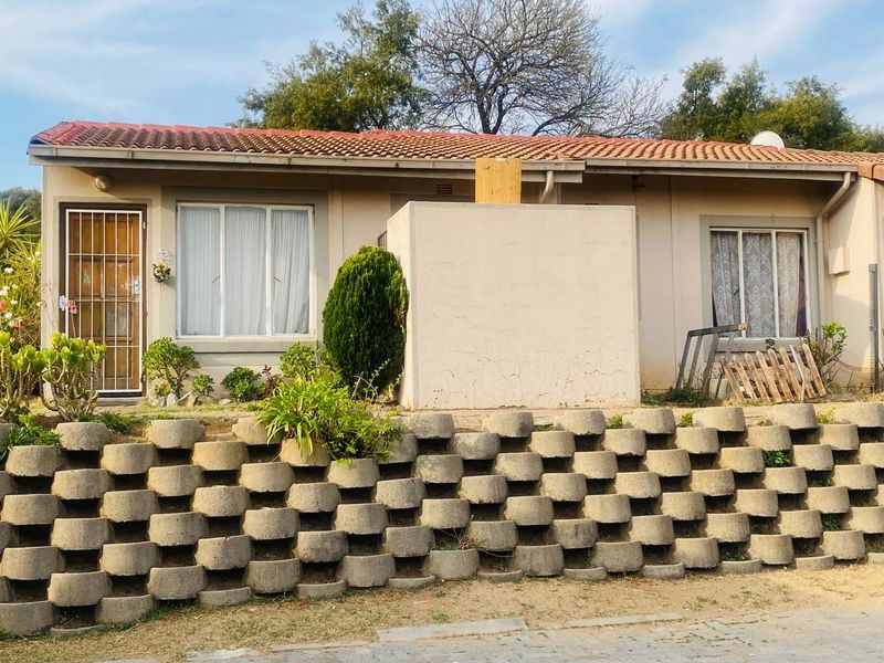 Front elevation of a single-story house featuring a terracotta tiled roof, security gate, and a prominent concrete block retaining wall.