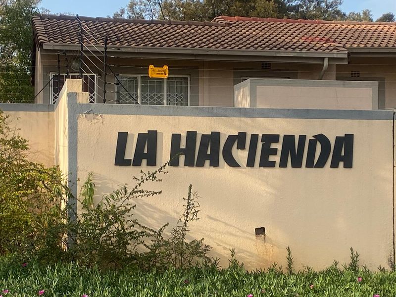 Front entrance view of La Hacienda complex featuring a concrete boundary wall with security fencing and tiled roof architecture.