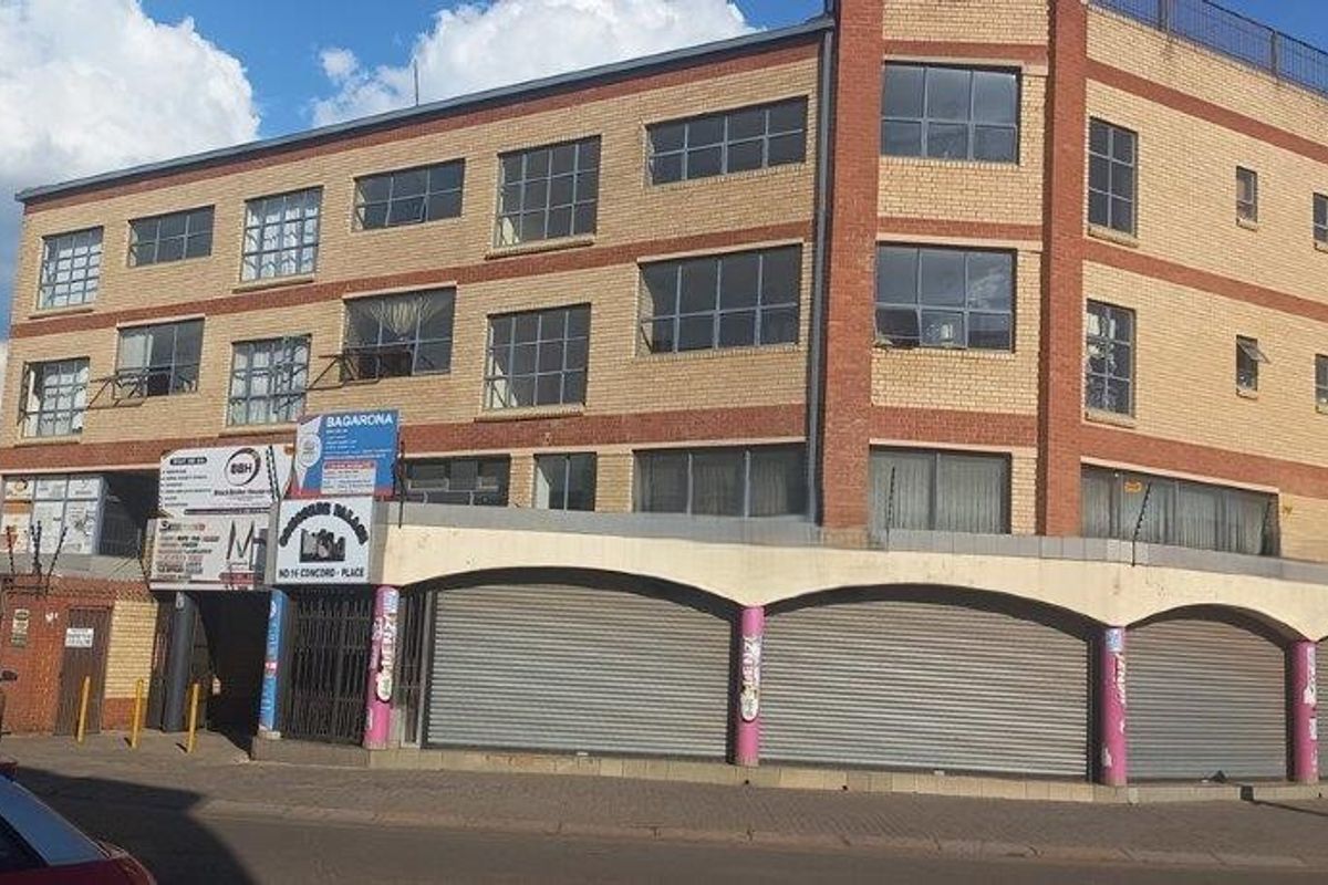 Multi-story commercial building featuring a brick facade, ground-level metal roller shutter doors, and security fencing along the perimeter.