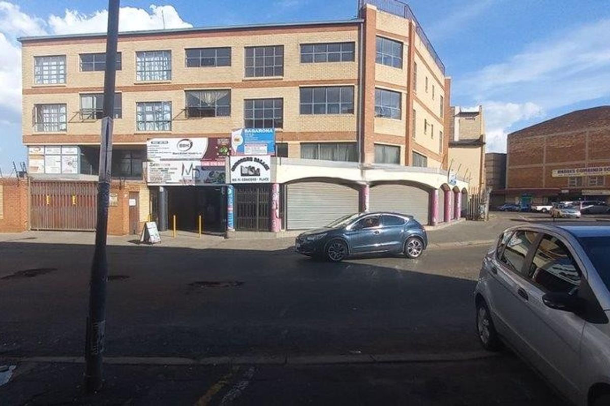 Street-level view of a multi-story commercial brick building featuring ground-floor retail units with security roller shutters and perimeter fencing.