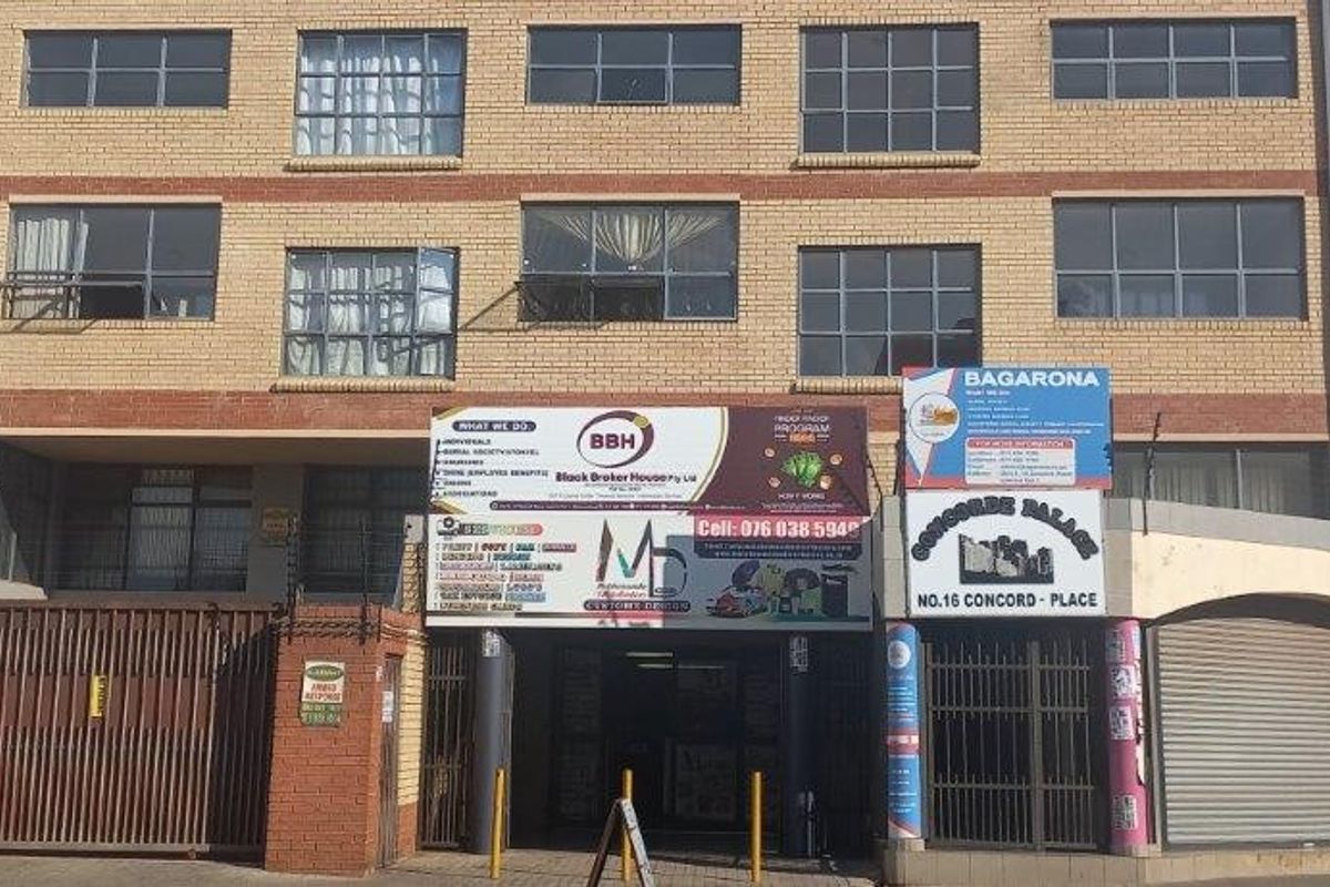 Front elevation of a multi-story commercial building featuring brick facade, aluminium windows, secure gated entrance, and ground-level roller shutter doors.