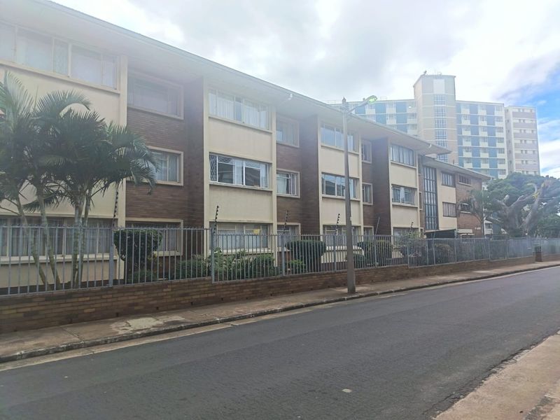 Street view of a multi-story apartment building featuring a brick facade, secure perimeter fencing, and an adjacent paved sidewalk.