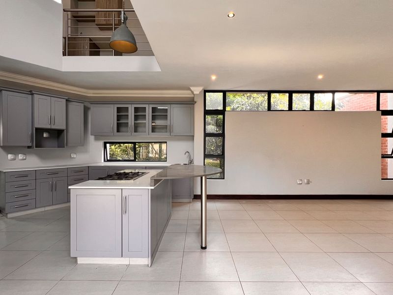 Spacious open-plan kitchen featuring modern grey cabinetry, a central island with a gas hob, and large windows, set within a bright, double-volume living area.