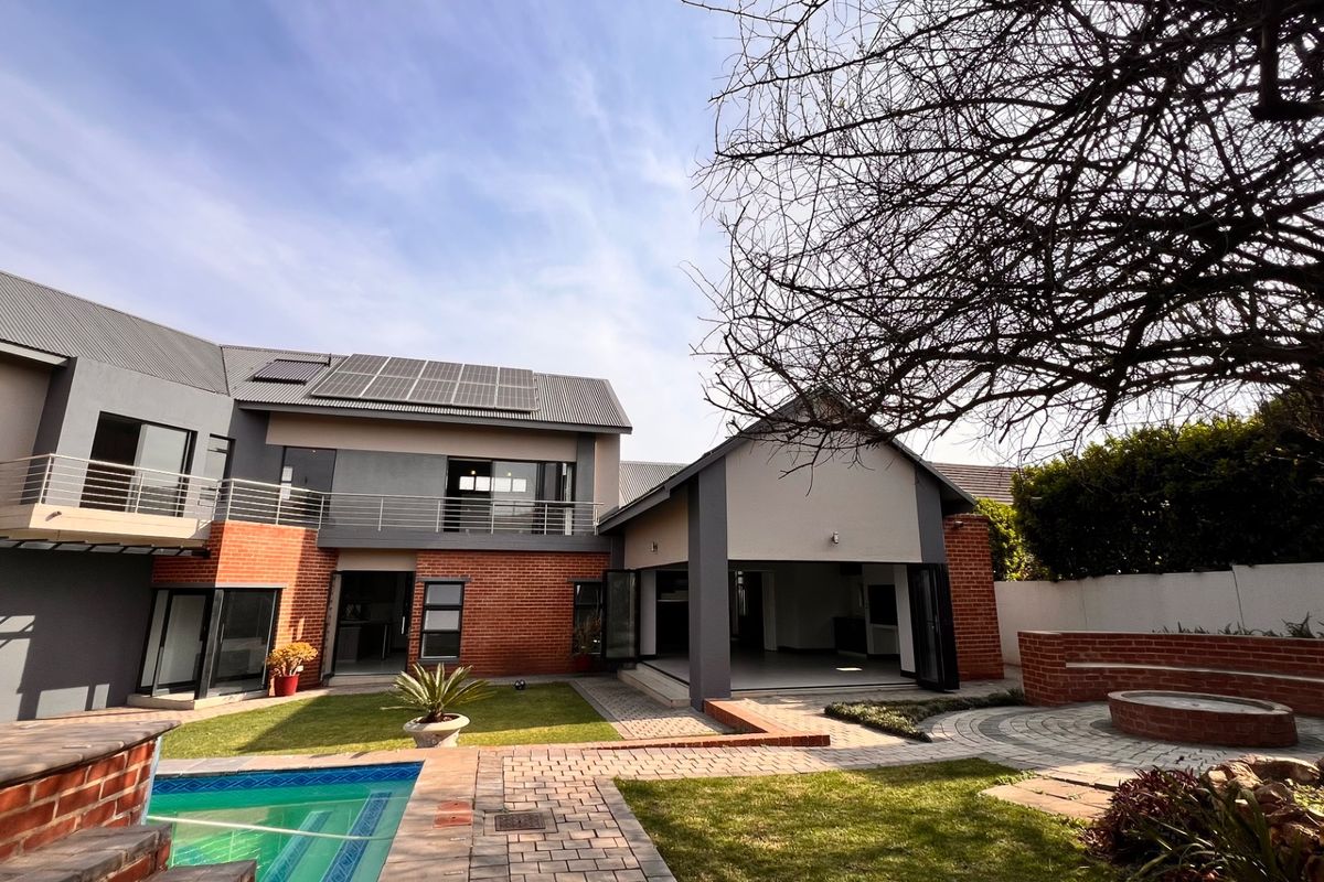 Spacious rear exterior featuring a swimming pool, paved patio with built-in seating, modern brick and render facade, balcony, and roof-mounted solar panels.