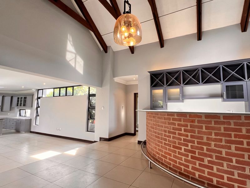 Spacious open-plan living area featuring high vaulted ceilings with exposed timber beams, a built-in brick bar, and a modern kitchen with tiled flooring.
