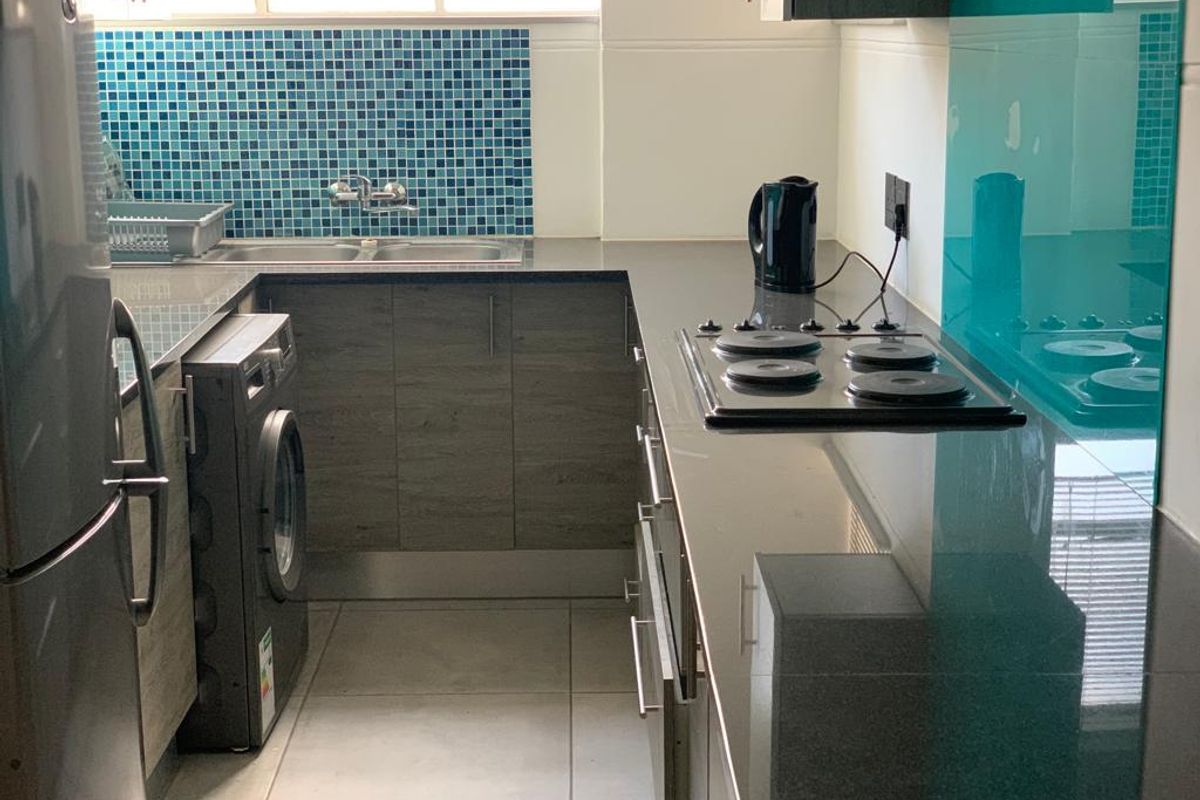 A compact, modern kitchen featuring dark wood-look cabinetry, stone countertops, a mosaic tile backsplash, and a built-in electric hob.