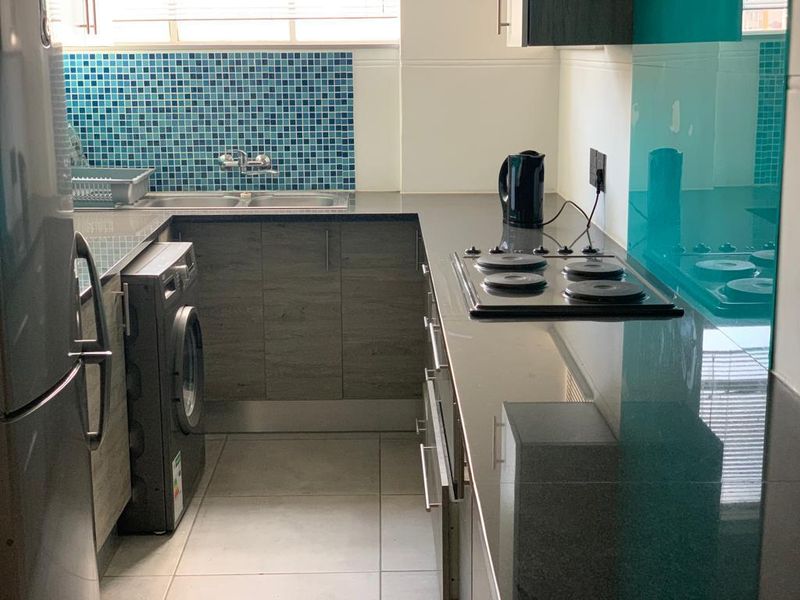 A compact, modern kitchen featuring dark wood-look cabinetry, stone countertops, a mosaic tile backsplash, and a built-in electric hob.