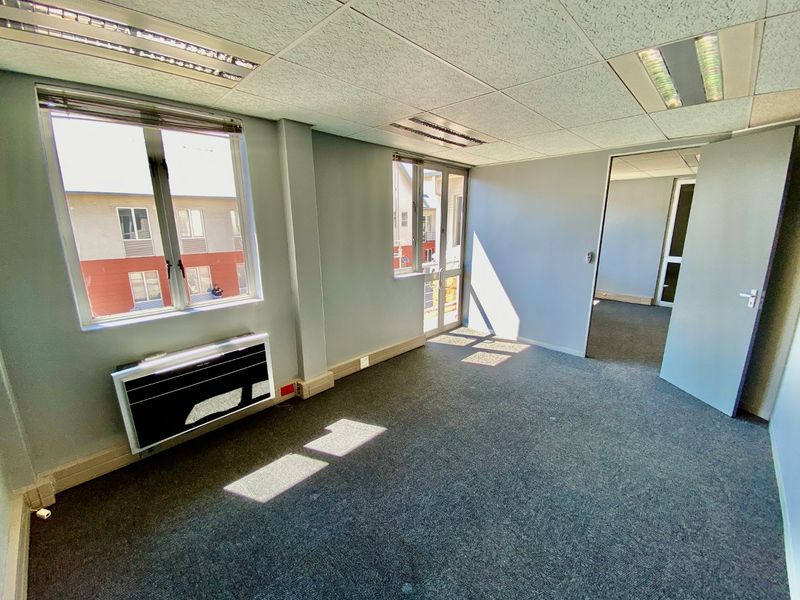 An empty commercial office space featuring carpeted flooring, a suspended ceiling with recessed lighting, and a wall-mounted air conditioning unit.