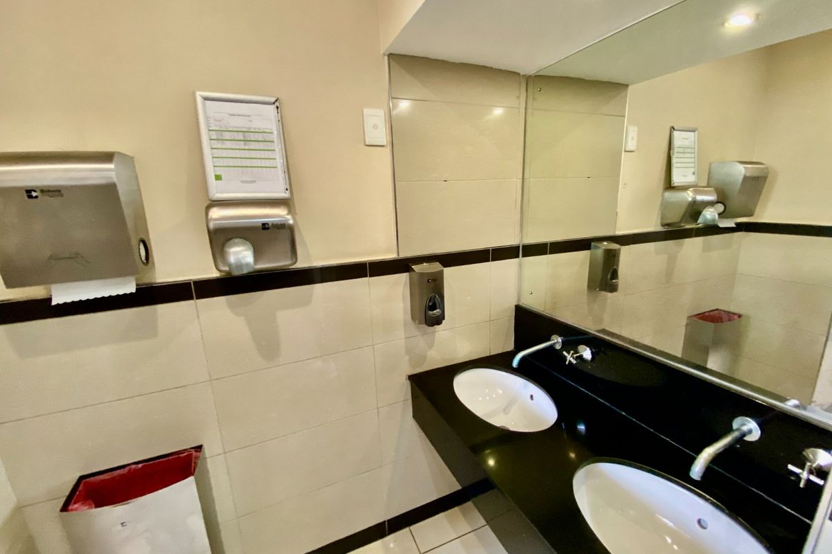 A clean commercial bathroom featuring a dual vanity with black countertops, large wall mirror, and wall-mounted utility dispensers.