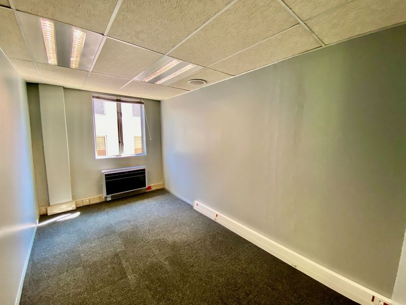 A vacant commercial office space featuring a suspended ceiling, recessed lighting, wall-mounted air conditioning, and carpeted flooring.