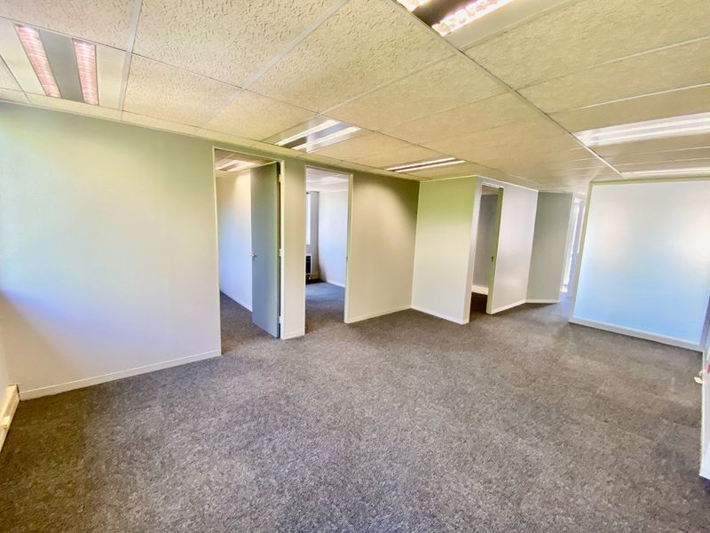 An open-plan commercial office space featuring a suspended ceiling with recessed lighting, grey carpeting, and multiple internal doorways leading to private office rooms.